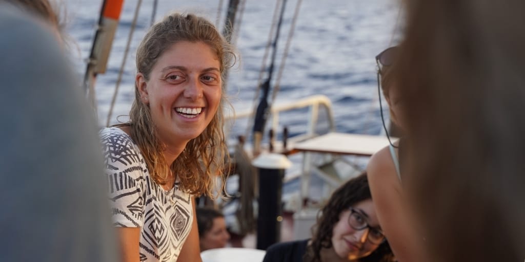 Mara is a young white woman with light coloured hair, picture on a sailing boat on the ocean. There are others on the sailing ship slightly out of shot.