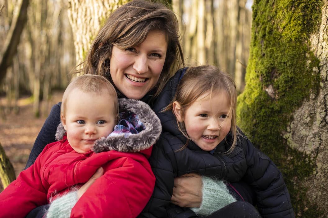 Picture shows Hanna squeezing two small children, they are all wrapped up in warm coats. In the background you can see blurred forest and mossy tree trunks. Hanna and both children are smiling. 