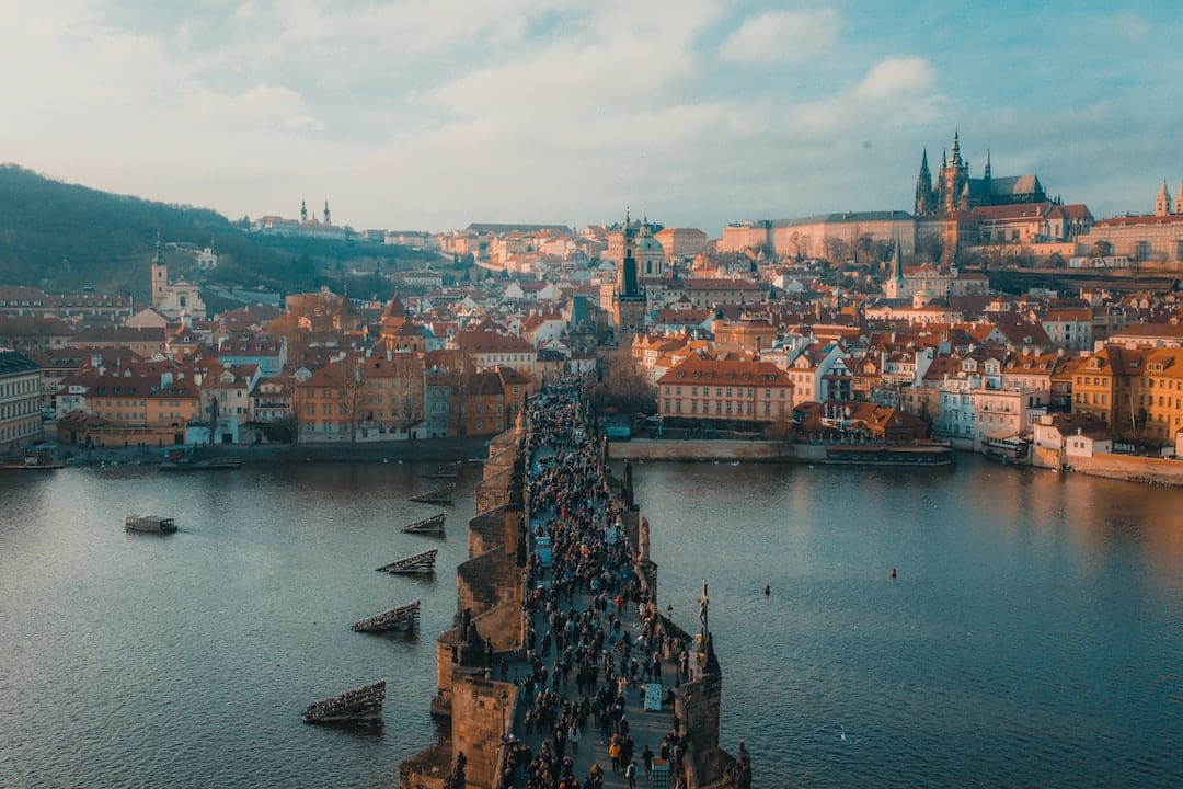 Image shows an aerial view of Prague, with people walking across the bridge over the river. The architecture in the background is a collection of terracotta roofs and spires.