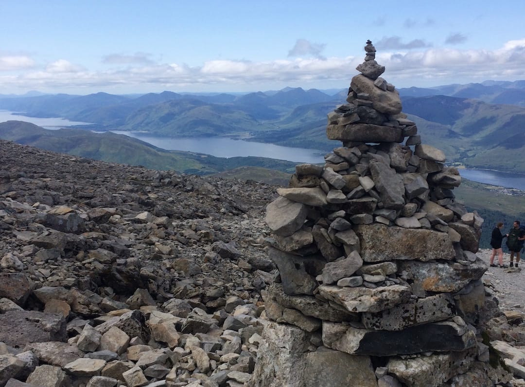 Image shows the view from near the summit of Ben Nevis on a clear day. There are a few clouds in the distance and miles of peaks. There is a cairn and lots of scree on Ben Nevis