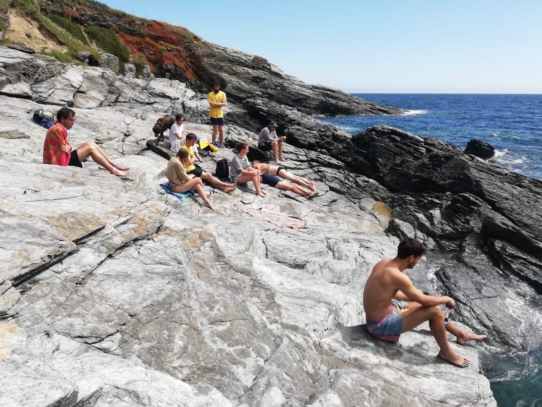 Image shows a group of people sitting on some rocks in the sun, next to the sea