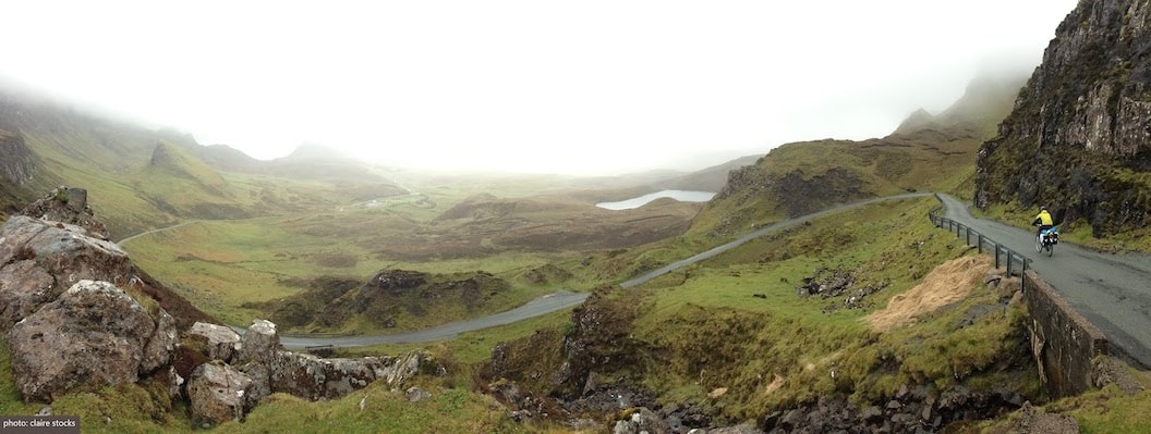 Picture shows a mountain-side road on the Isle of Skye. The terrain is green, rocky and marshy, and low fog is covering the peaks of the hills. There is a small body of water in the distance. 