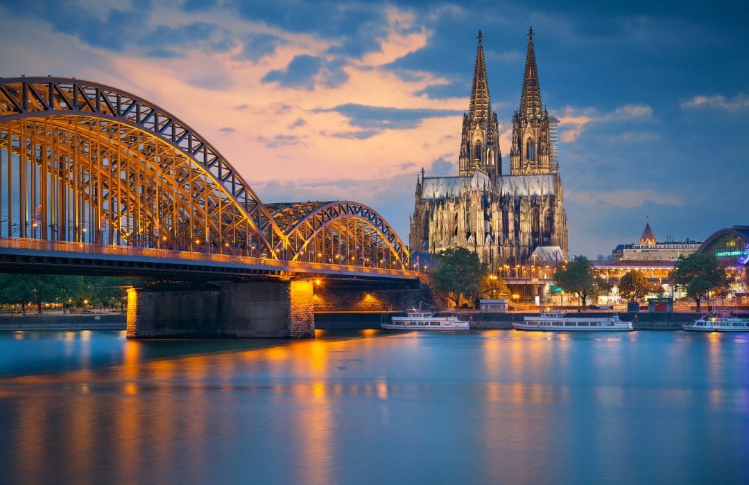 Picture shows a river, a bridge and a lit up cathedral in the city of Cologne. It is night time and the city is lit up with blue and yellow lights. There are illuminated trees in the foreground.