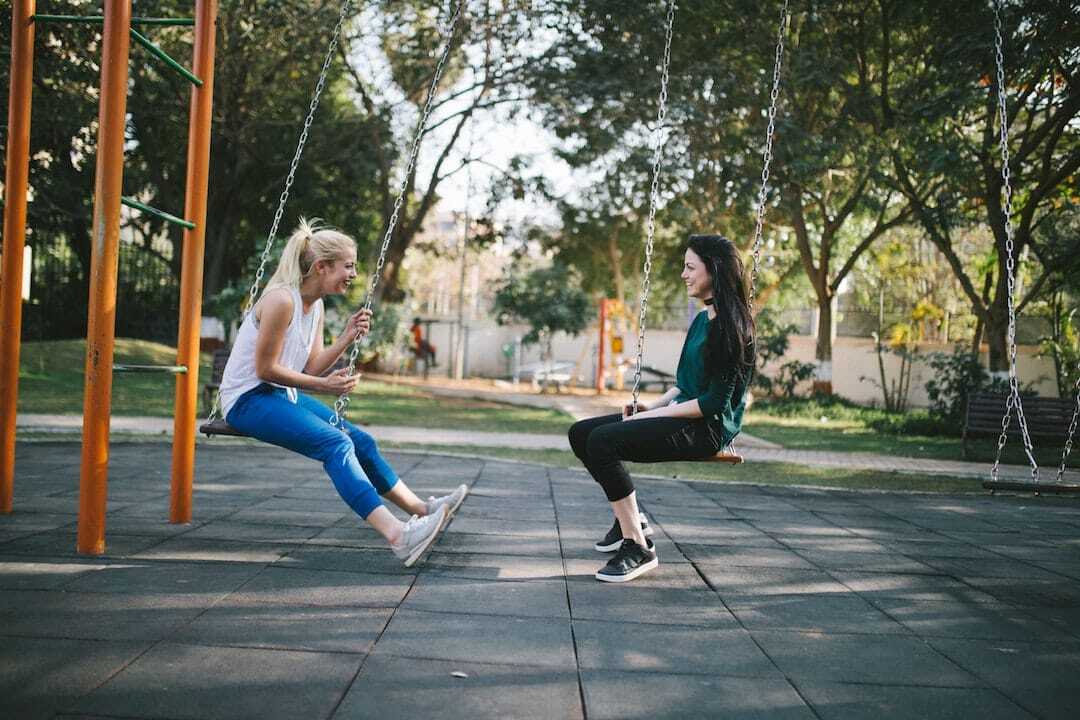 Image shows two women sitting on swings talking and laughing.