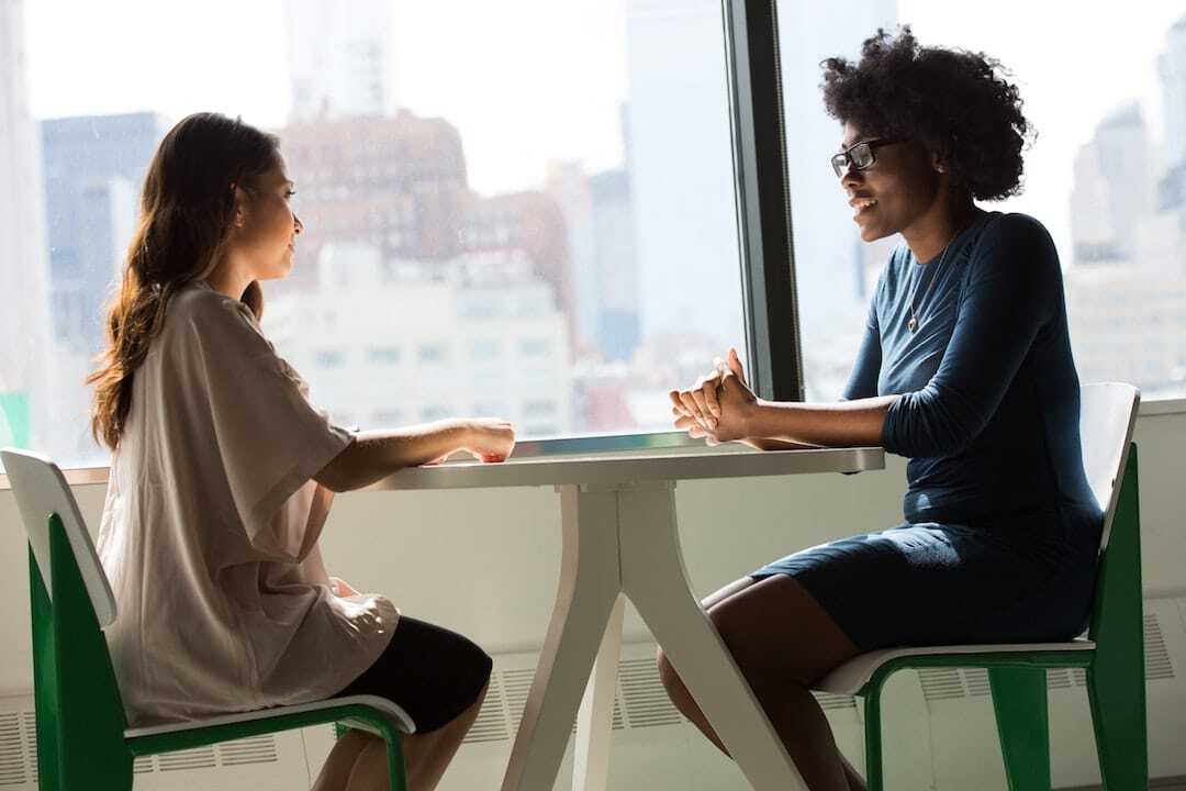 Image shows two women sitting beside a table talking.