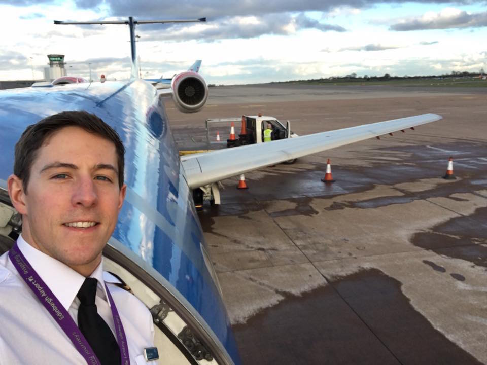 Picture shows dan taking a selfie out the door of a stationary grounded plane. He's smiling and behind him is a runway and a cloudy sky. 