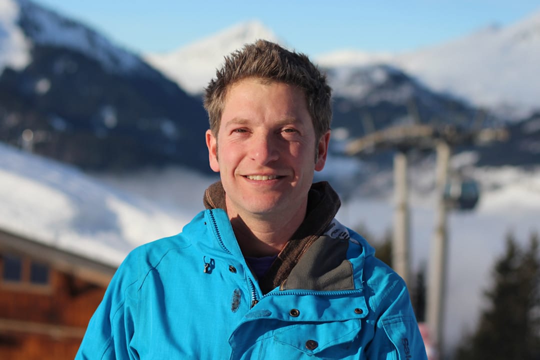 Picture shows Daniel smiling at the camera wearing a bright blue ski coat. In the background is a blurred wood chalet, along a snowy and sunny mountain view. 