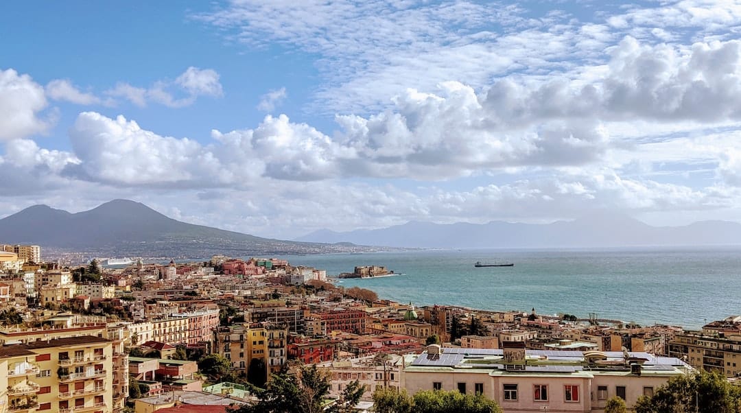 Image shows the bay of Naples. There is a mountain in the background and the many colourful buildings of Naples in the foreground.