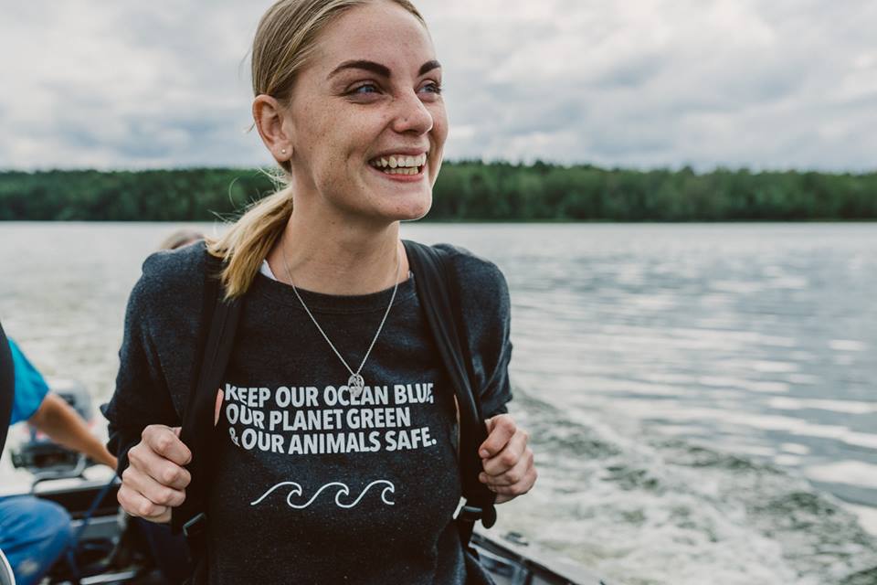 Evelina is smiling widely whilst being driven in a small boat. She is wearing a T-shirt that reads "Keep our oceans blue, our planet green and our animals safe". The are blurred trees in the background and an overcast sky. 