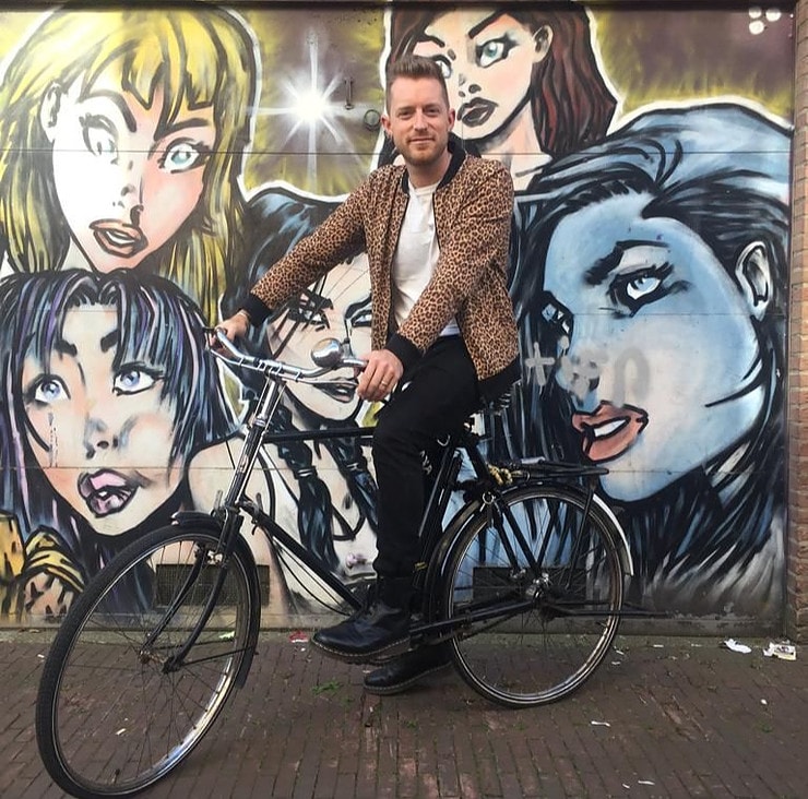 Picture shows Gavin sitting on a black bicycle. He is smiling and wearing a leopard-print jacket. Behind him is a wall of graffiti. 