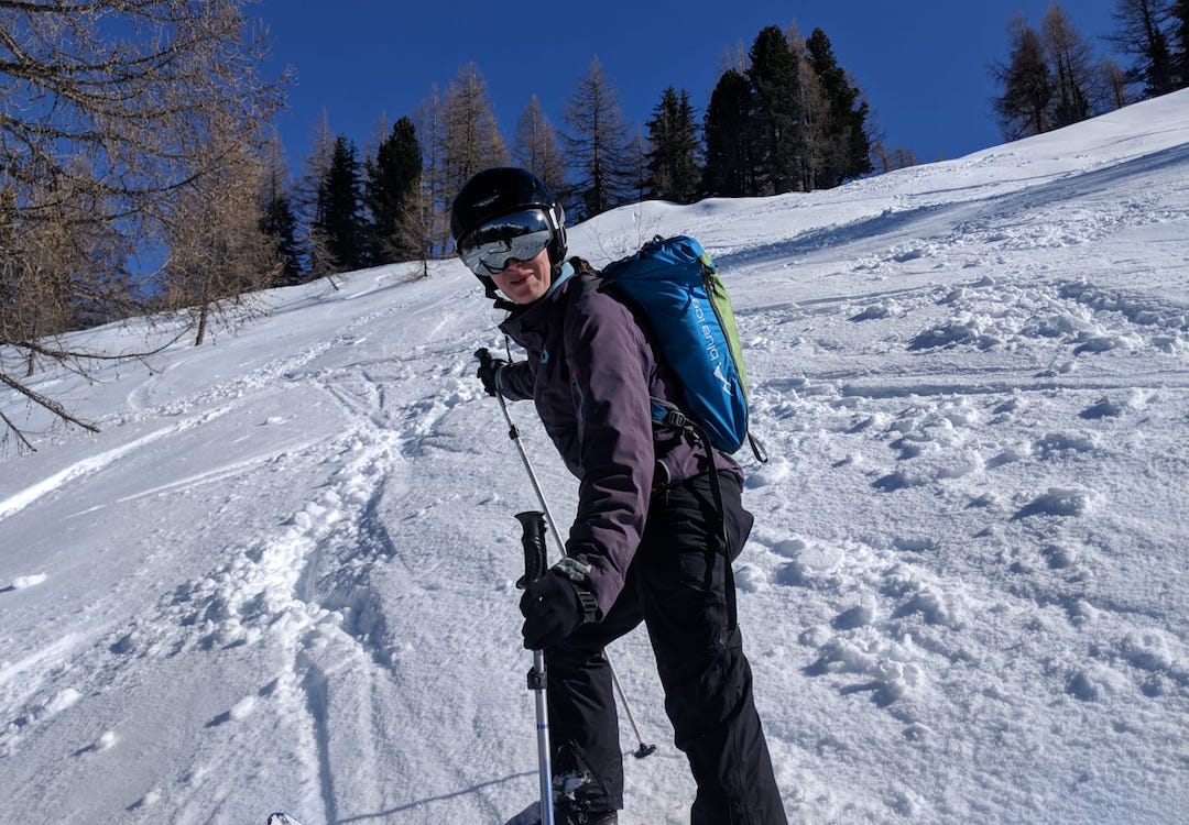 Picture shows skier facing the camera. She is wearing full ski gear including mirrored sunglasses and helmet. Behind them is and expanse of white snow with pine trees in the background. The sky is deep blue. 