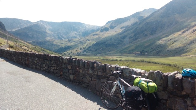 Picture shows a low stone wall with Helena's bike resting against it. Behind the wall are green grassy fields and rocky tall hill ranges. 