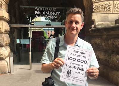 Picture shows Iain holding a piece of paper with the Flight Free 2020 pledge on it. He is standing outside the entrance to the Bristol Museum and Art Gallery, and is smiling. 