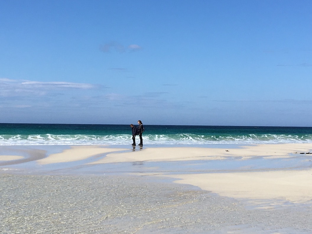 Picture shows two figures walking along a sandy beach with the tide very far out. The sea looks deep blue and turquoise and the sky is clear blue with few clouds. The sand is pale yellow and there is shallow pools and streams running through it. 