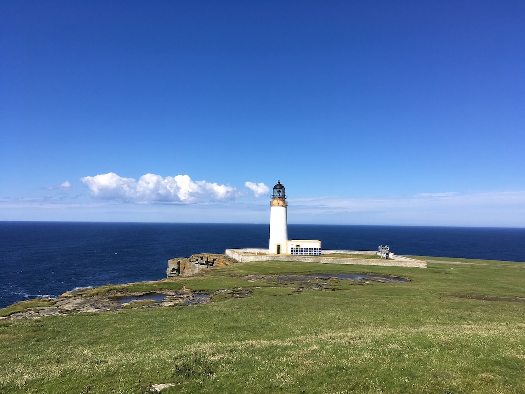 Picture shows a white lighthouse on the edge of a green grassy cliff. The sea behind looks a deep indigo and the sky is clear except for some small fluffy clouds. 