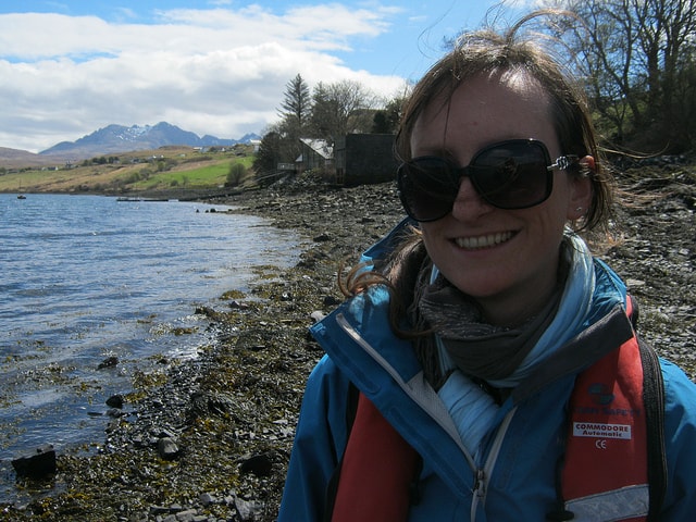 Anna stands next to a lake in the Isle of Skye. Behind her are two modern houses, green fields and faraway mountains. The sky is blue with large clouds. 