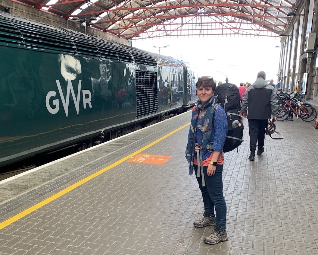 Picture shows Jools next to a stationary train in Penzance station. She's smiling and carrying a large rucksack. 
