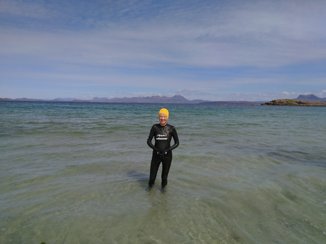 Kate stands in a wetsuit in the sea up to ankle height. She is wearing a bright yellow swimming cap and smiling. The sea looks clear and in the distance are craggy mountain tops out to sea. 