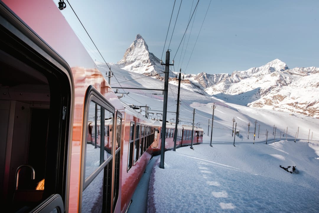 Pitcure shows a photo taken out of a mountain train window. The side of the train is red and snowy peaks are visible close behind the train. It is surrounded by snow and the sky is clear blue. 