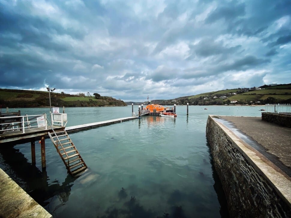 Picture shows a small pier with an orange lifeboat and a few smaller boats at the end of it. The sea is a muted turquoise green and the sky is moody with dark clouds. There are green hills in the background. 