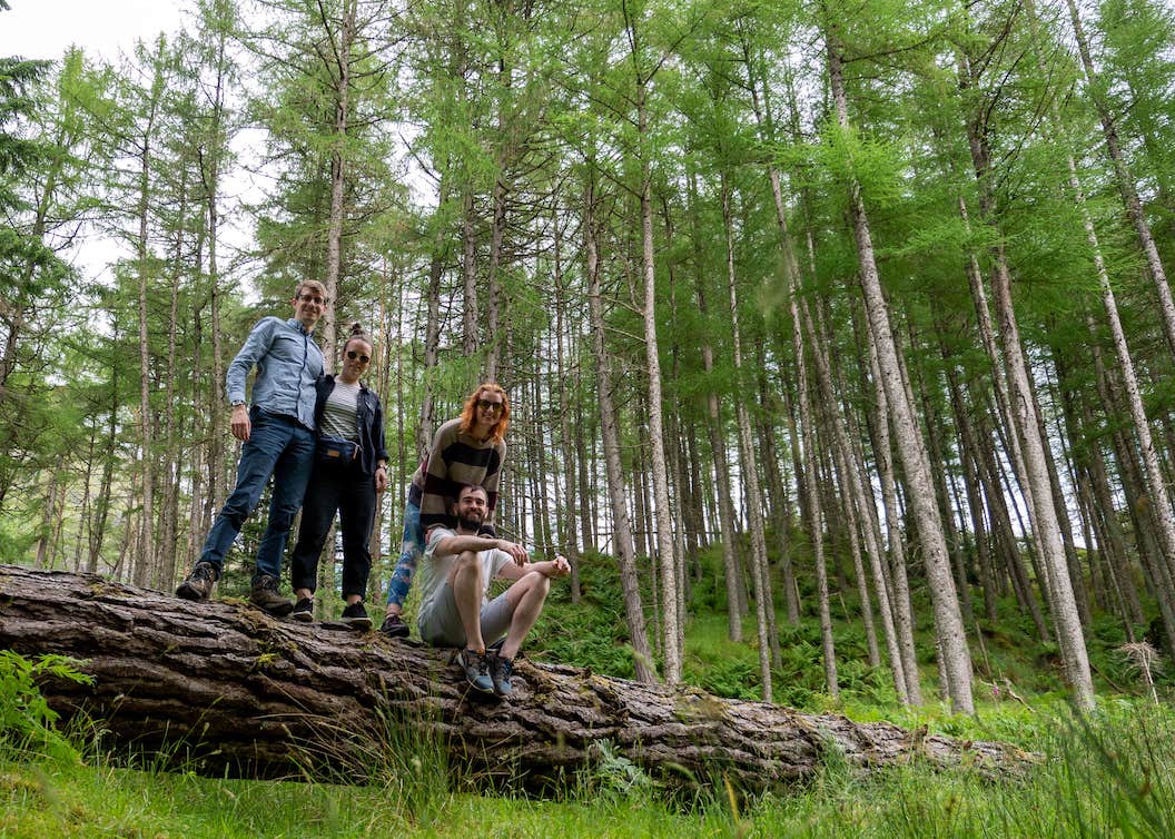 Picture shows one person sitting on a log and three others standing. They are smiling to pose for the picture. Behind the log is a forest of thin trees. 
