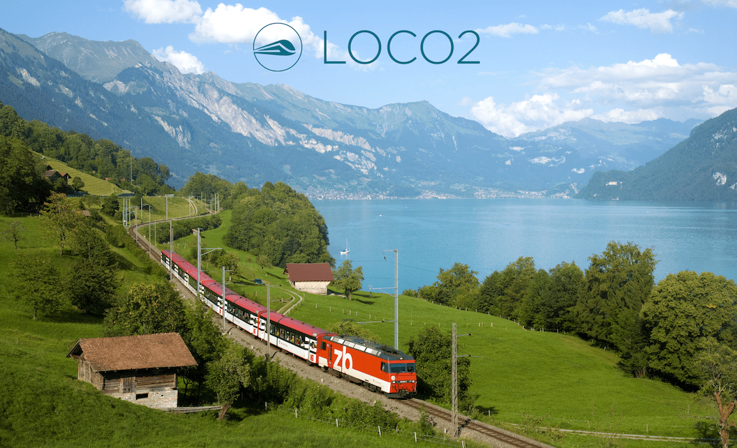 Picture shows a red and white train travelling along a track next to a lake in a wide andlush valley. There are mountain peaks in the background and the sky is blue with large fluffy clouds. 