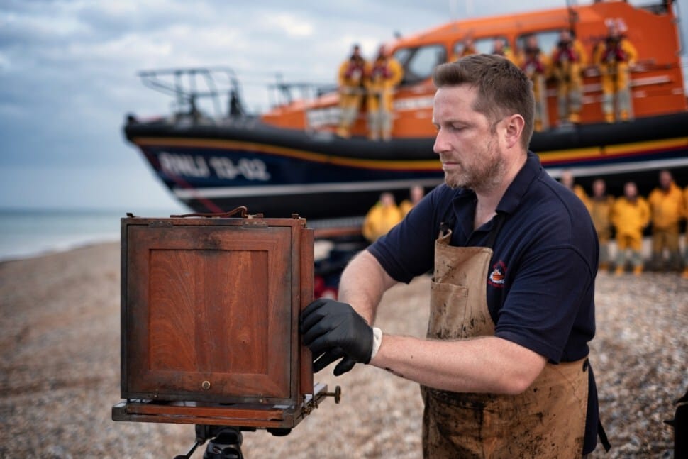 Picture shows Jack Lowe operating an old camera on a pebble beach in front of a large lifeboat with its crew. The lifeboat and the crew are blurry and Jack is wearing brown dungarees and black gloves. 