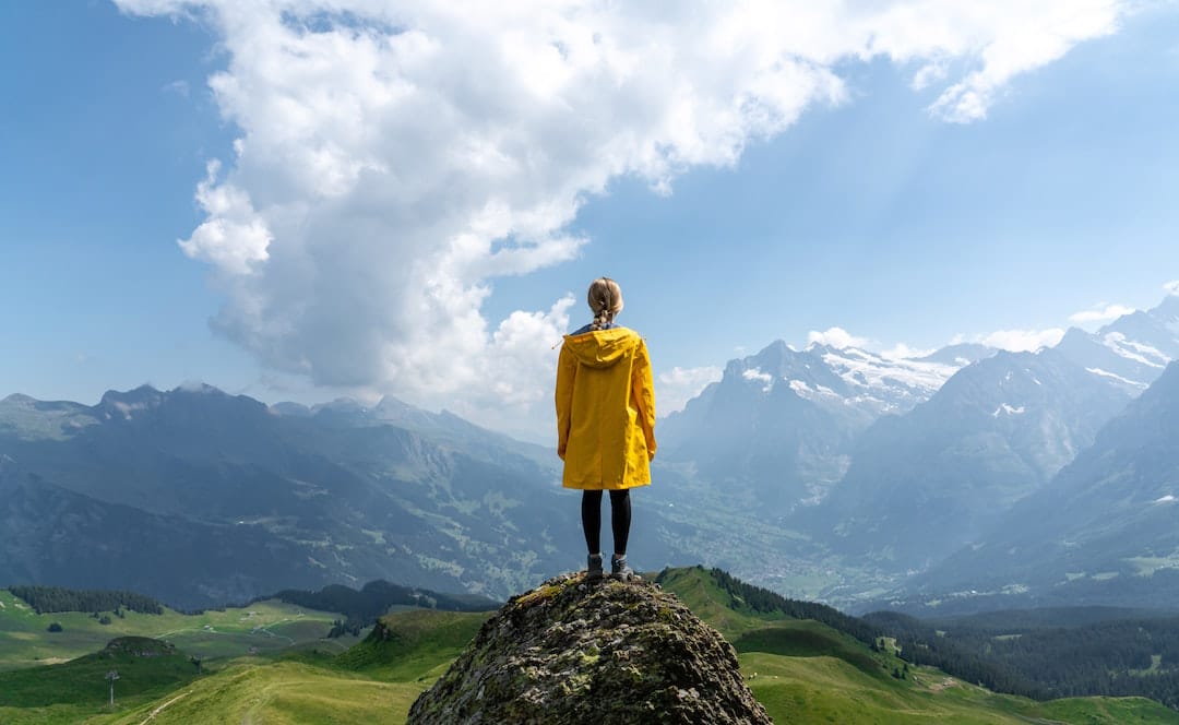 Picture shows a women in a yellow raincoat standing on the top of a craggy hill top. She has her back to the camera and in front of her is a mountain range and green rolling hills. The sky is blue and cloudy. 