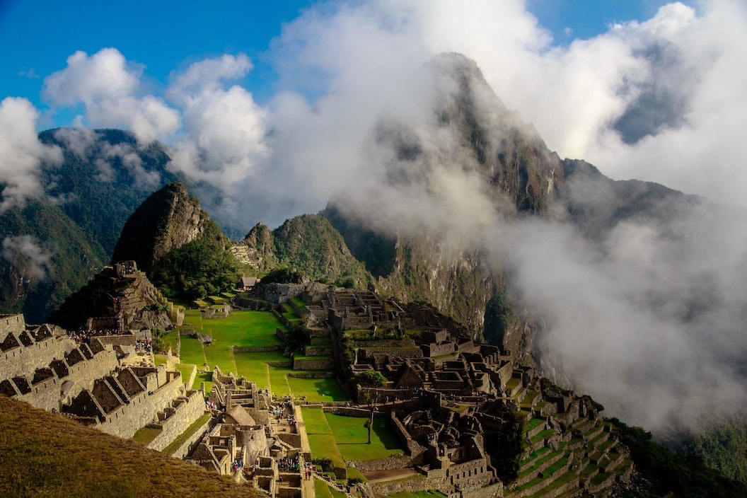 Picture shows Machu Picchu with its steep tree covered peaks and expansive stone structures and levels. Clouds are covering taller peaks that surround Machu Picchu in the background. 