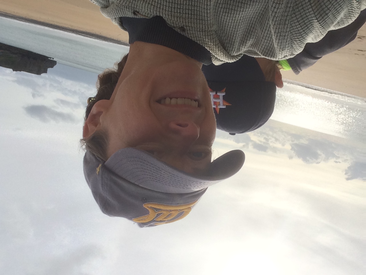 Picture shows Maggie taking a selfie, smiling and wearing a cap. Her son ducks his face behind her shoulder. Behind them is a view of the sea and pale sunny sky. The surface of the sea is glittering. 