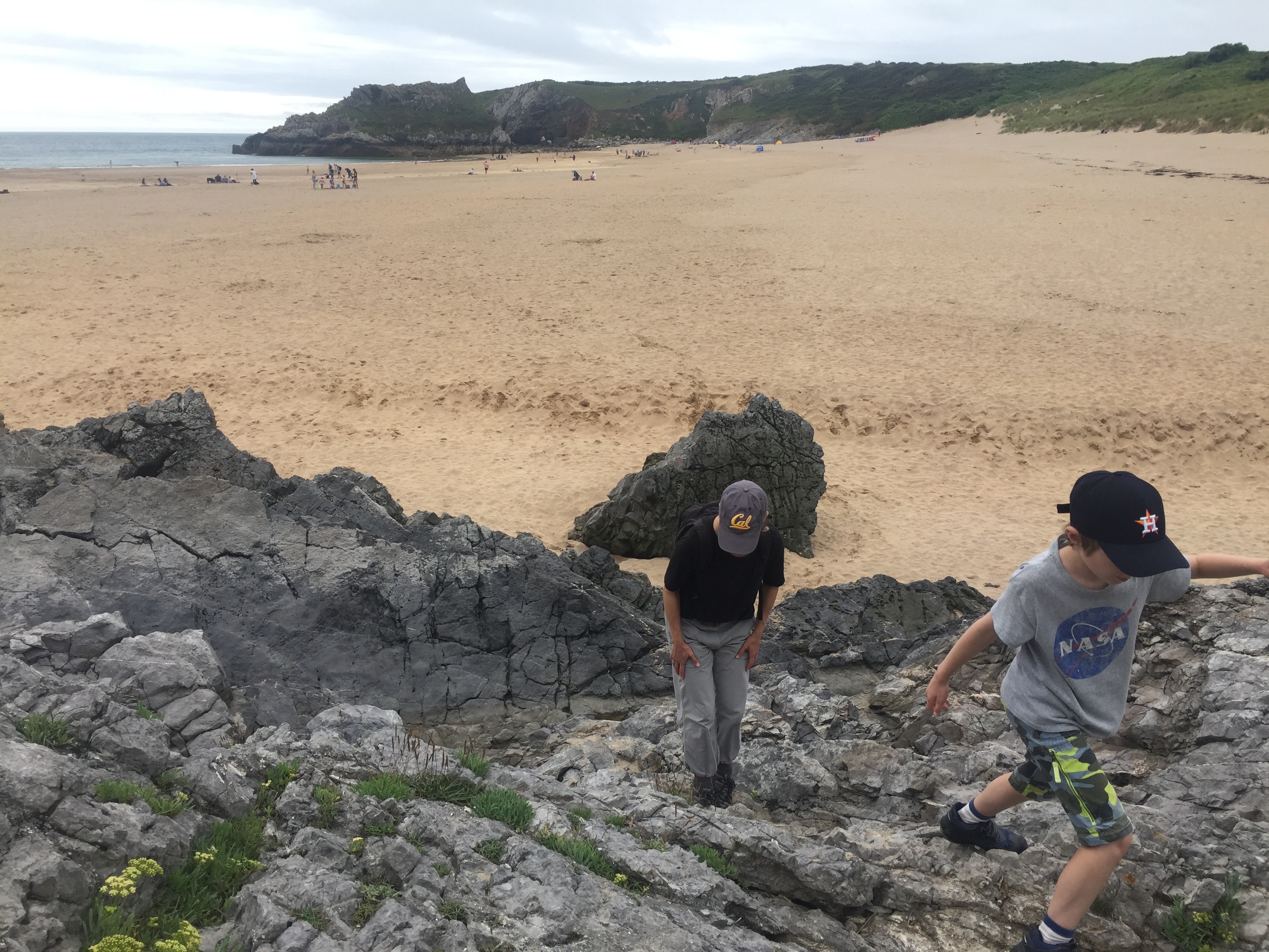 Picture shows Maggie and her son scrambling over grey rocks on the beach. Both are wearing caps and looking down. You can see hills in the background. 