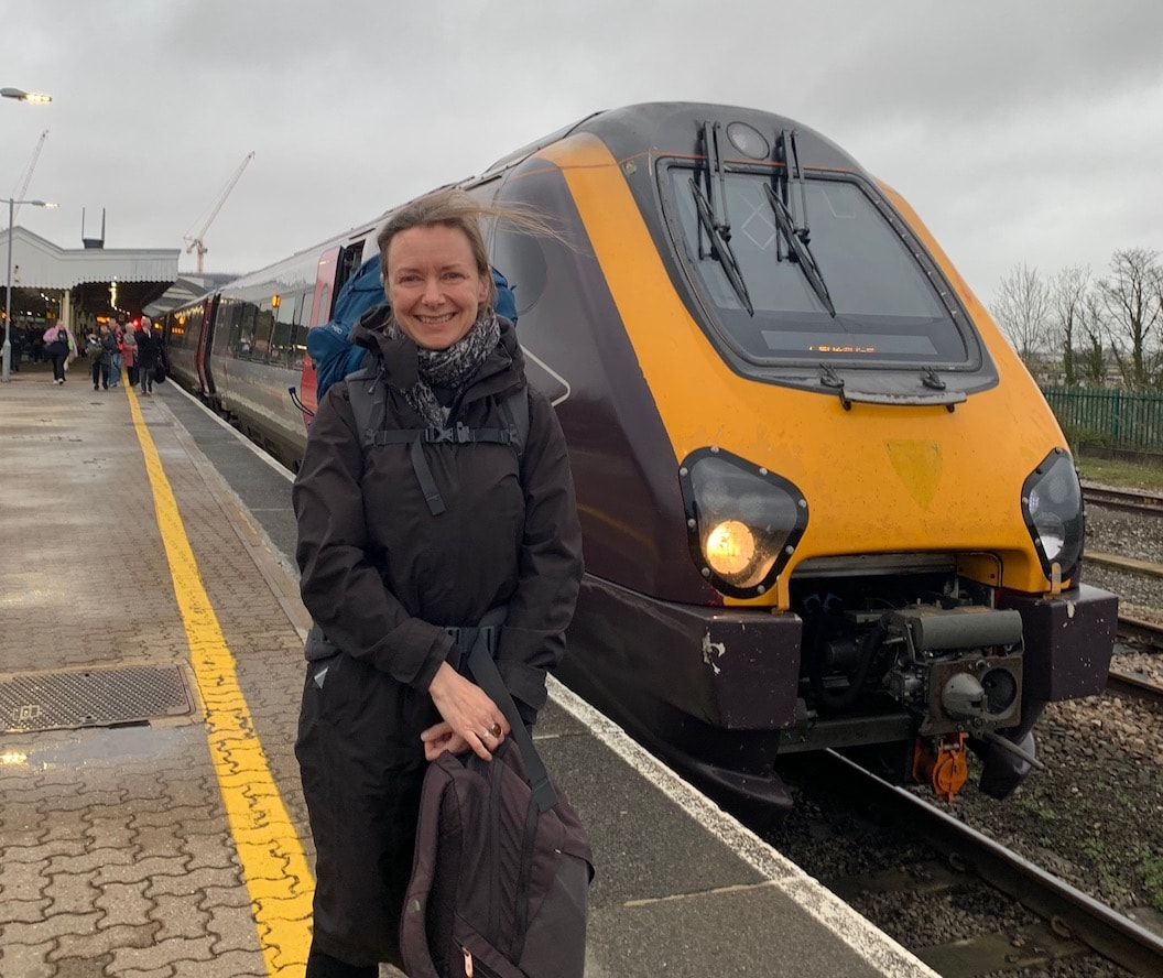 Picture shows Maggie standing next to a stationary train at the station. She is smiling and wearing a coat and scarf, and carrying a large backpack. 