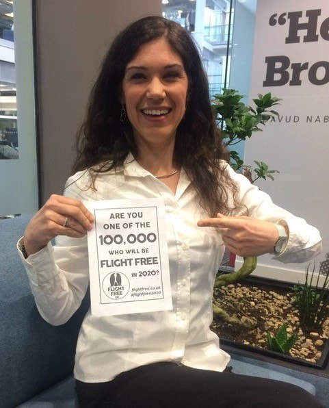 Picture shows Milena holding a piece of paper with the Flight Free 2020 pledge on it. She is sat on a sofa in an office environment, she is wearing a white shirt and smiling. 