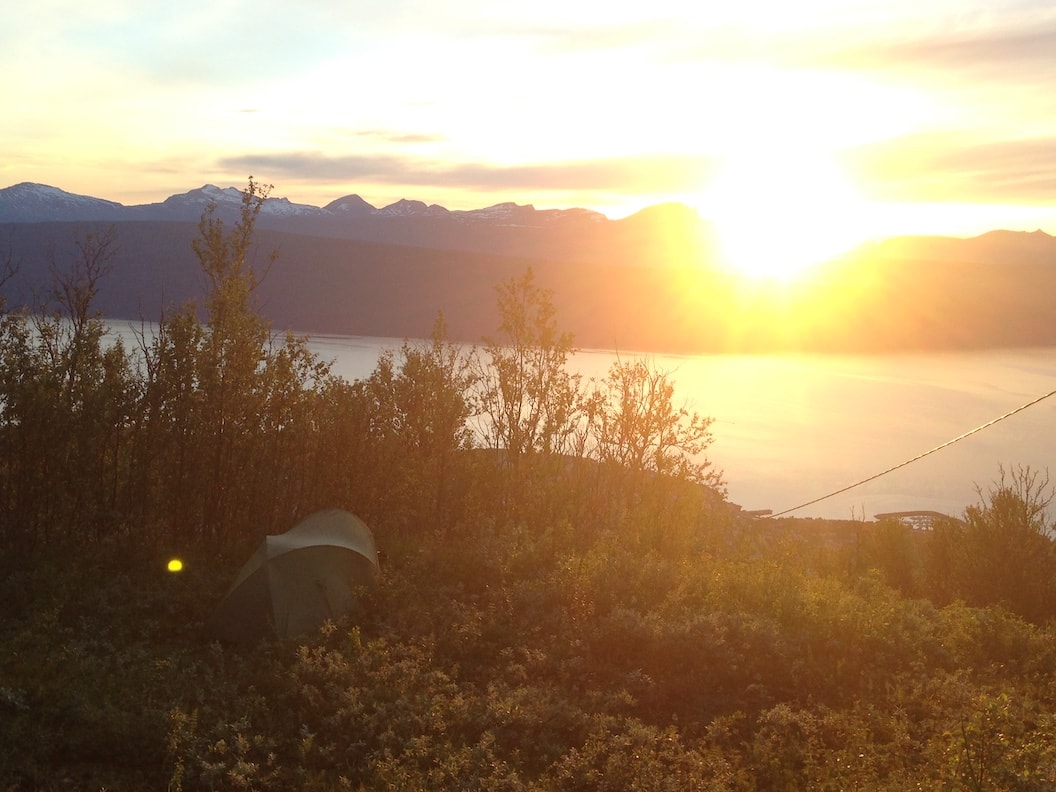 Picture shows a sun setting over a lake. In the foreground are shrubs and trees and a tiny tent. 
