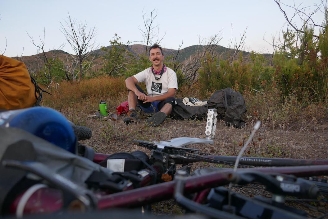 Picture shows Mike sat down on the ground in front of some shrubs. It appeard to be dusk and his bike is laying down in front of him. He's smiling. 