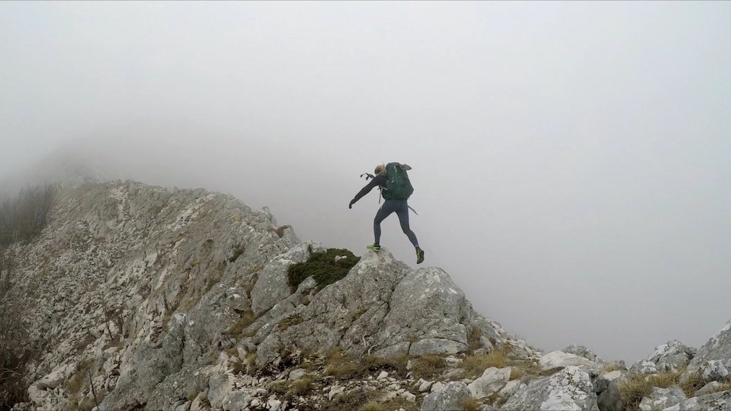 Picture shows Rosie jumping energetically over craggy and mossy grey rocks. The view behind her is completely obscured by a wall of fog and nothing else is visible. She's wearing sporty clothes and carrying  large rucksack. 