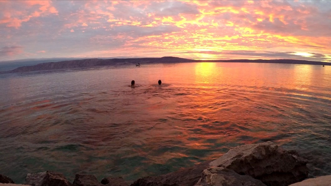 Picture shows Rosie and Mike swimming in a clear body of water. The sun is setting over the horizon and the sky looks bright orange and pink. In the distance you can see the silhouette of some low hills. 