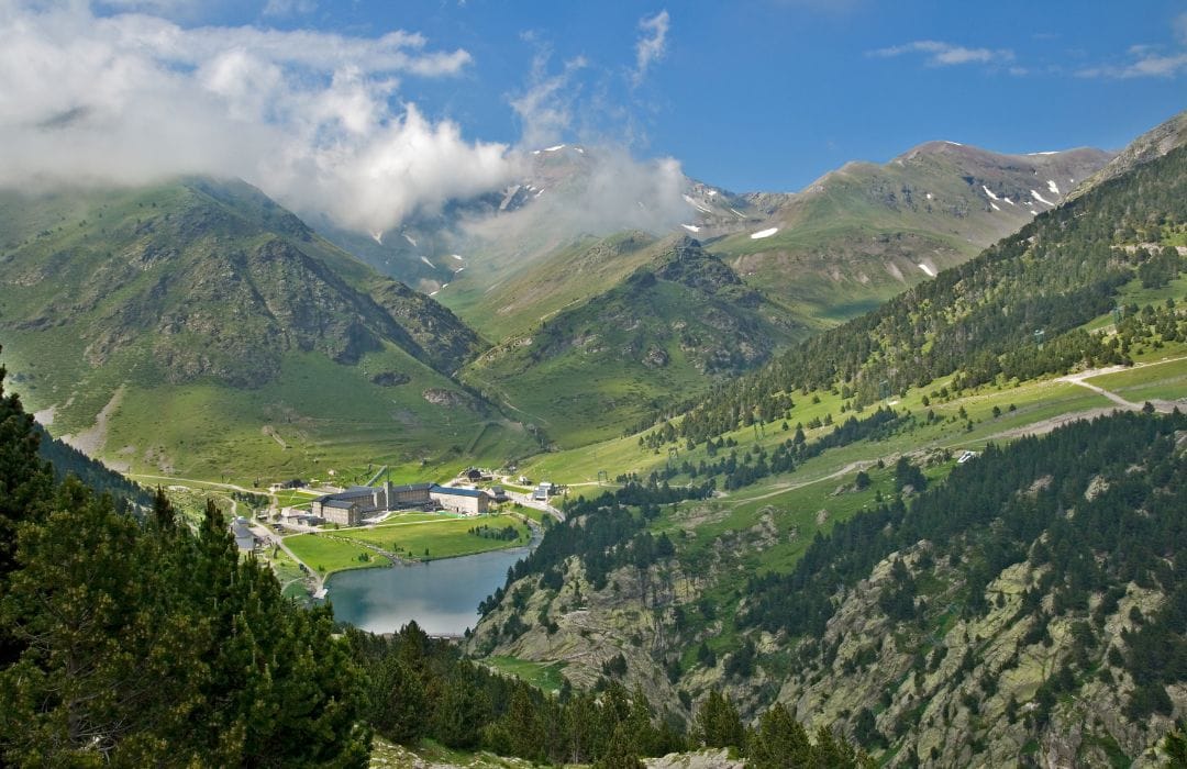 Picture shows the Pyrenees mountains. There is a lush green grassy valley with trees lining the sides. The hills of the valley lead up to the Pyrenees mountain range and the peaks are covered in clouds. The sky is blue.