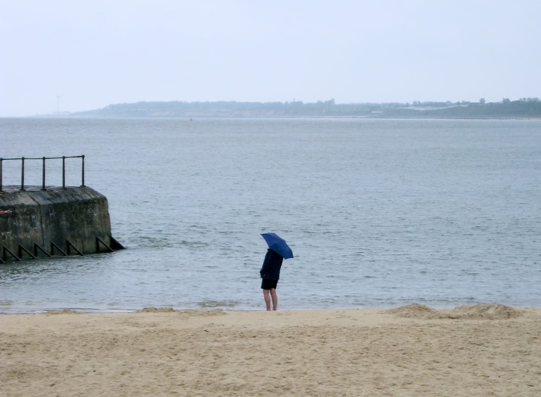 A person stands on a deserted beach holding a blue umbrella