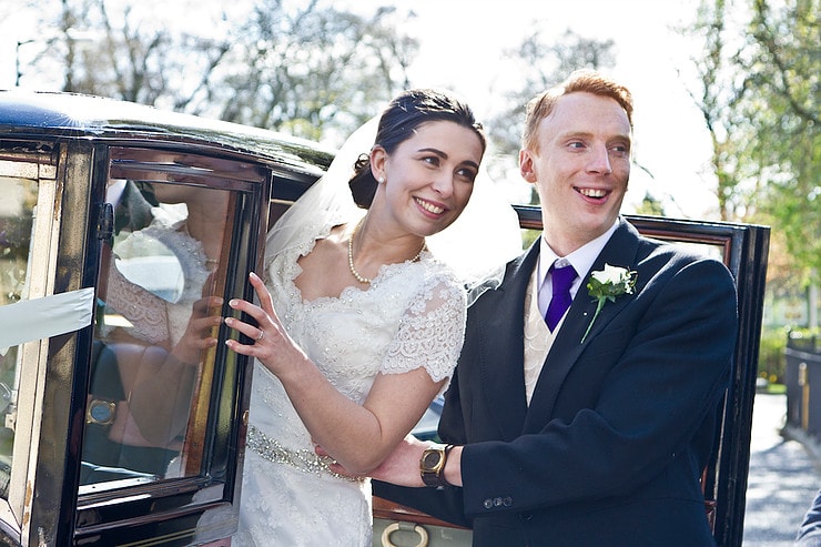 Picture shows Rebecca and Marc on their wedding day. Marc is helping Rebecca out of an old fashioned car. She is wearing a classic wedding dress with lace detail and a veil, he is wearing a dark blue suit with a purple tie. 