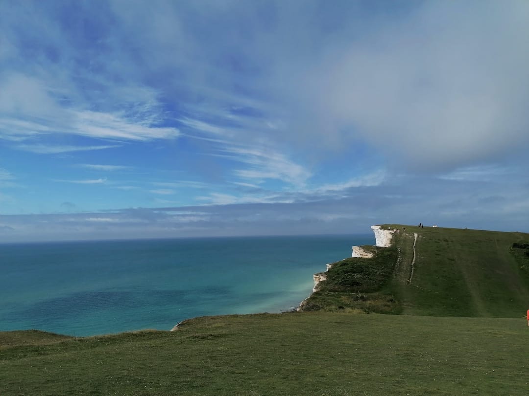 Image shows the South Downs Way: a grassy footpath on the top of chalk cliffs, with a blue sea and whispy clouds in the sky