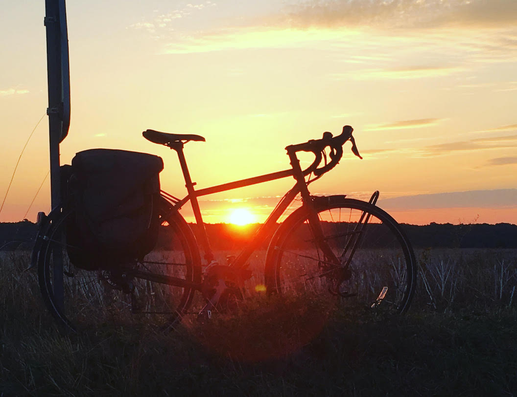 Picture shows a bike from ground level. Through the frame of the bike you can see the sunsetting in the background. 