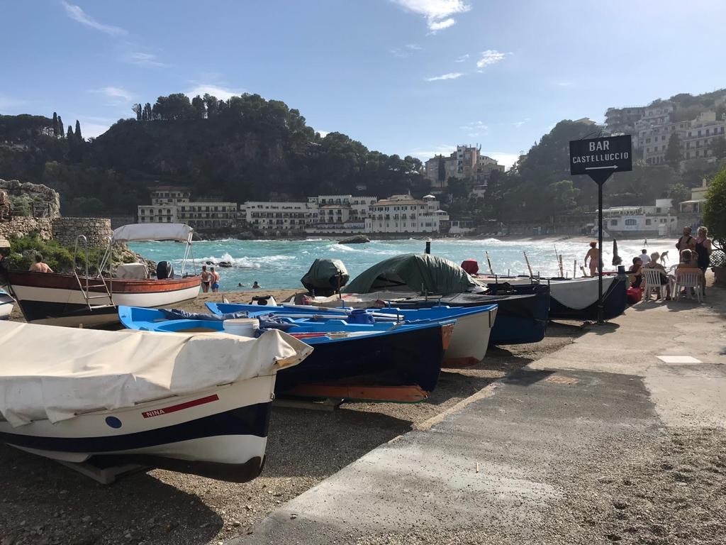 Picture shows boats moored on the beach of a harbour. The waves behind are bright blue and choppy. On the other side of the water are old-fashioned hotels. 