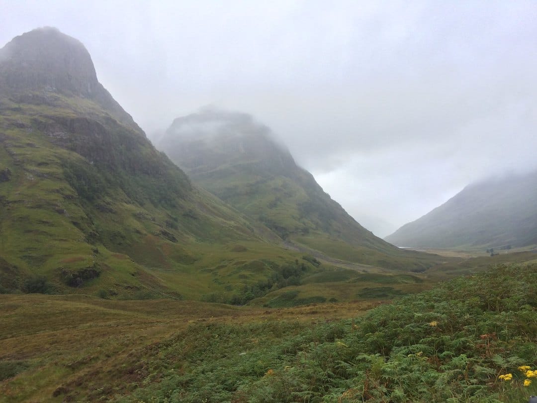Pictures shows the Three Sisters in Glencoe: very steep mountainous peaks covered in green ferns and purple heather, seen on a very rainy and cloudy day