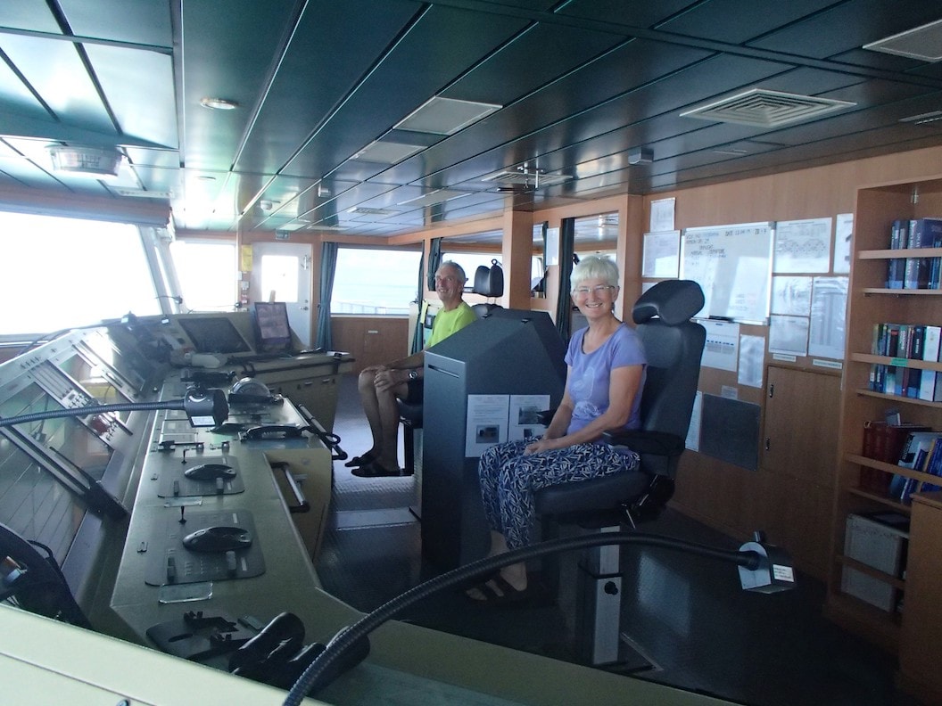 Peter and Christine sit in two chairs at the bridge of the ship. They are both smiling. 