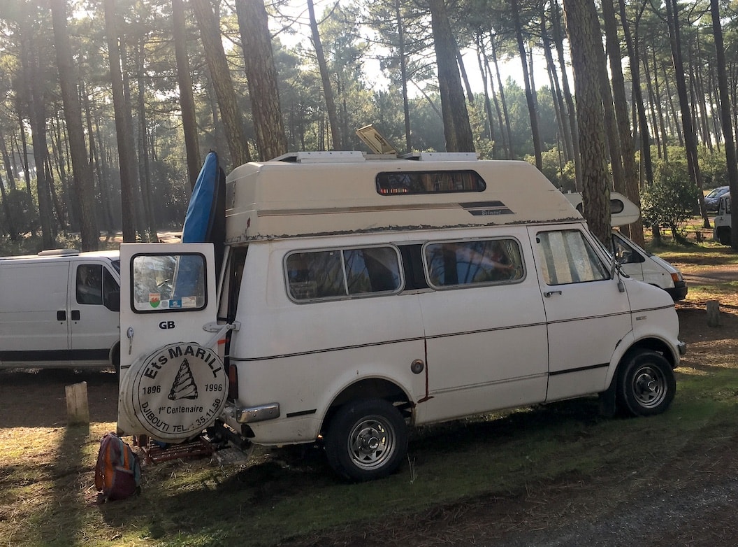 A white campervan has its back door open. It is parked in the clearing of a forest of tall pine trees. Shade is dappled on the forest floor.