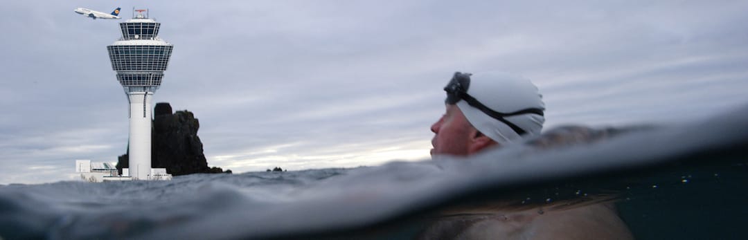 Picture shows a swimmer in the sea, with an airport watch-tower and aeroplane on a rock. 